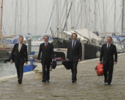Four male musicians wearing dark suits, walking on a stone pier toward the viewer. The men are holding string instrument cases, and sailboats are in the background.