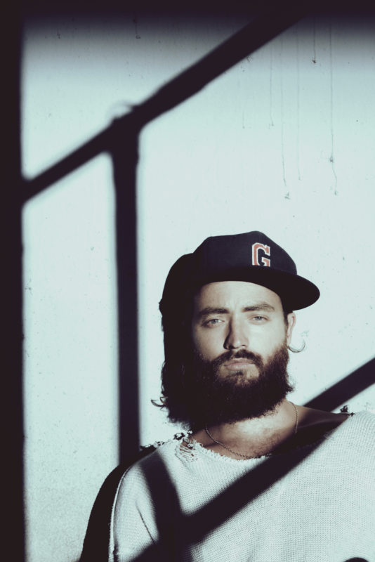 Man with a beard and a baseball cap wearing a tattered white t-shirt, looking directly at the viewer stands against a white wall with the shadow of a stair railing across the wall and his chest.