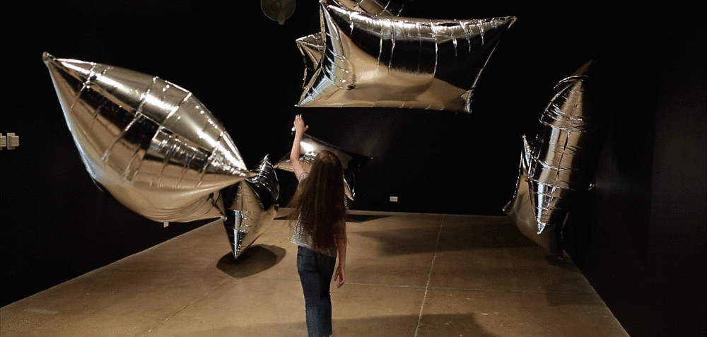 A child reaches a hand out to touch one of the large silver foil balloons that comprises the silver clouds art installation.