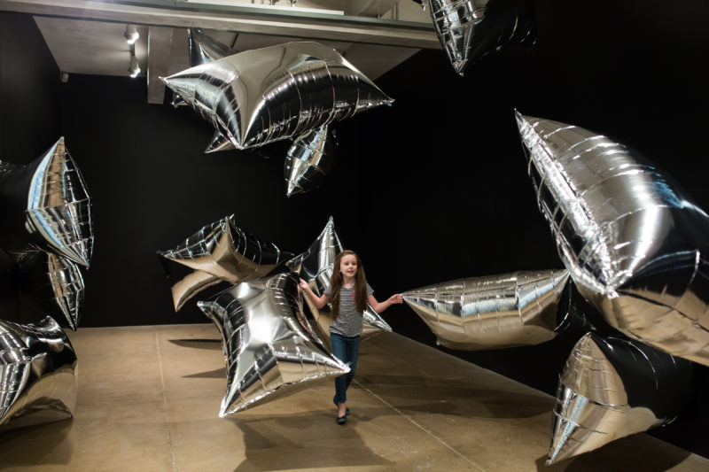A young girl with long brown hair wearing a black and white striped shirt and skinny jeans interacts with large, rectangular balloons in The Andy Warhol museum’s Silver Clouds exhibit.