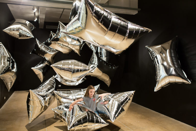 A girl with long, brown hair, wearing a black and white striped shirt and jeans interacts with large, silver, rectangular balloons in The Andy Warhol Museum’s Silver Clouds gallery.