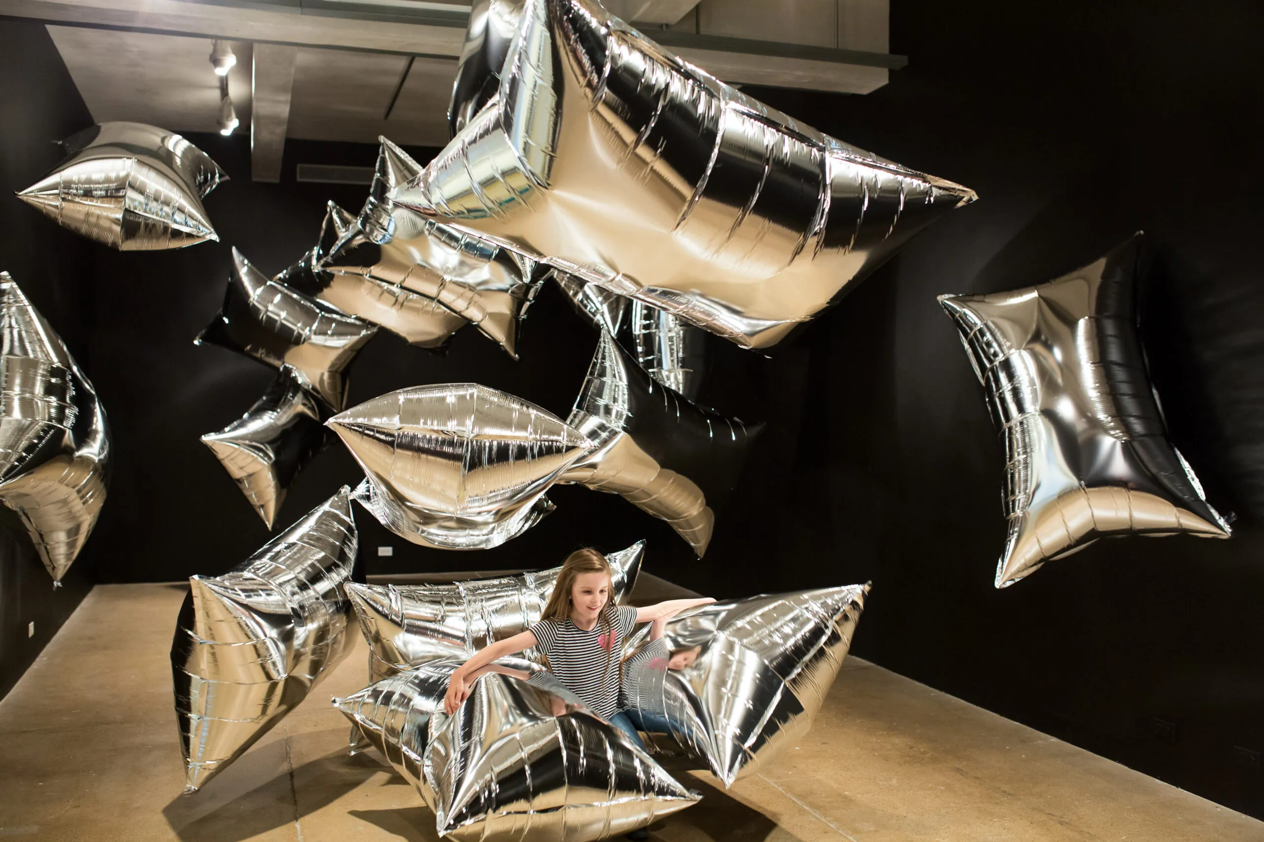 A girl with long, brown hair, wearing a black and white striped shirt and jeans interacts with large, silver, rectangular balloons in The Andy Warhol Museum’s "Silver Clouds" gallery.