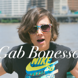 Gab Bonesso wearing sunglasses and a Nike t-shirt looks directly at the viewer, mouth open. In the background is a river and buildings on the opposite river bank.
