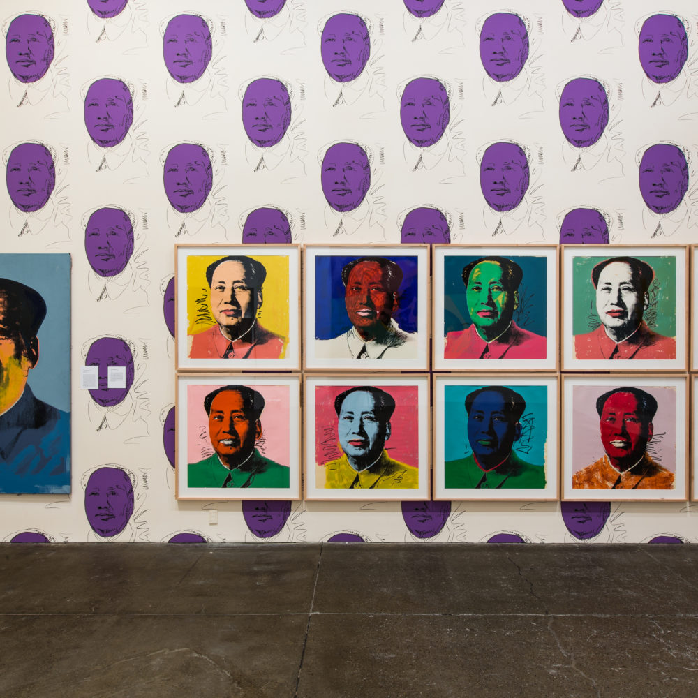 Eleven Mao portraits in different colors hanging on a wall wallpapered with purple Mao portraits.