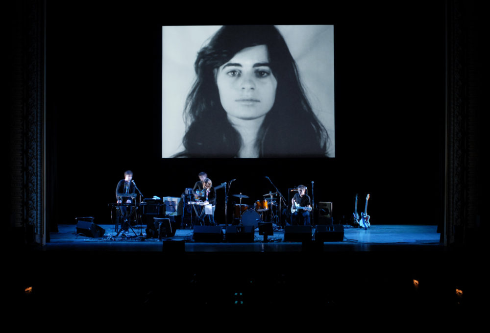 A band performs on stage in front of a black and white photograph of a woman with long dark hair that is projected on the wall behind them.