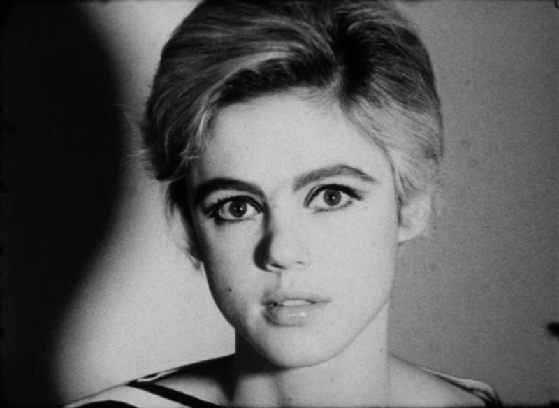 A young woman with short blonde hair gazes into the camera, her plump lips slightly parted. She sits against a light background, and her shadow falls to the left side of the image.