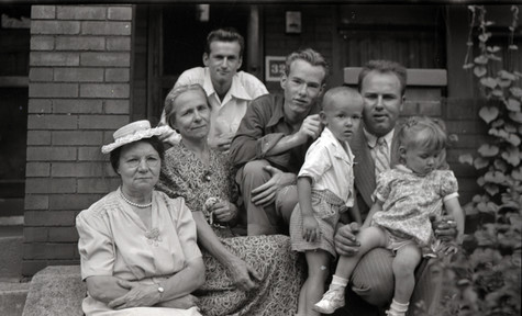 A black and white photograph of the Warhol family sitting on the front stoop of a brick house. Andy Warhol, a young man, sits between two of his brothers. On the right, one of his brother is holding two young children. Two women sit on the left.