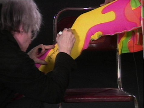 Wearing a black sweatshirt and turning away from the camera, Andy Warhol signs someone's leg on an episode of Andy Warhol's Fifteen Minutes. The leg is clad in yellow pants with large, organic blocks of other bright colors, including hot pink, red, and green.