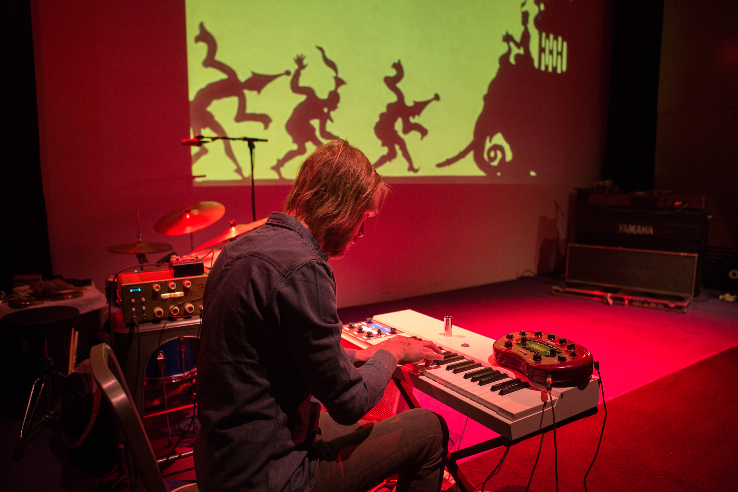 A man with shoulder-length blond hair sits at a keyboard on a stage bathed in red light. In the background, silhouettes of figures wearing hats and an elephant are projected against a yellow-green background.