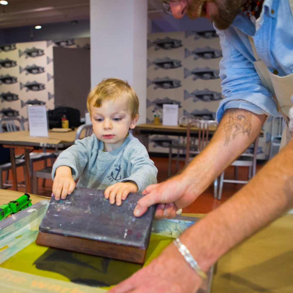 An artist educator wearing a blue button-down shirt with his sleeves rolled to his elbows and a beige apron helps a toddler with blonde hair make a screen print in the Andy Warhol Museum's Factory studio.