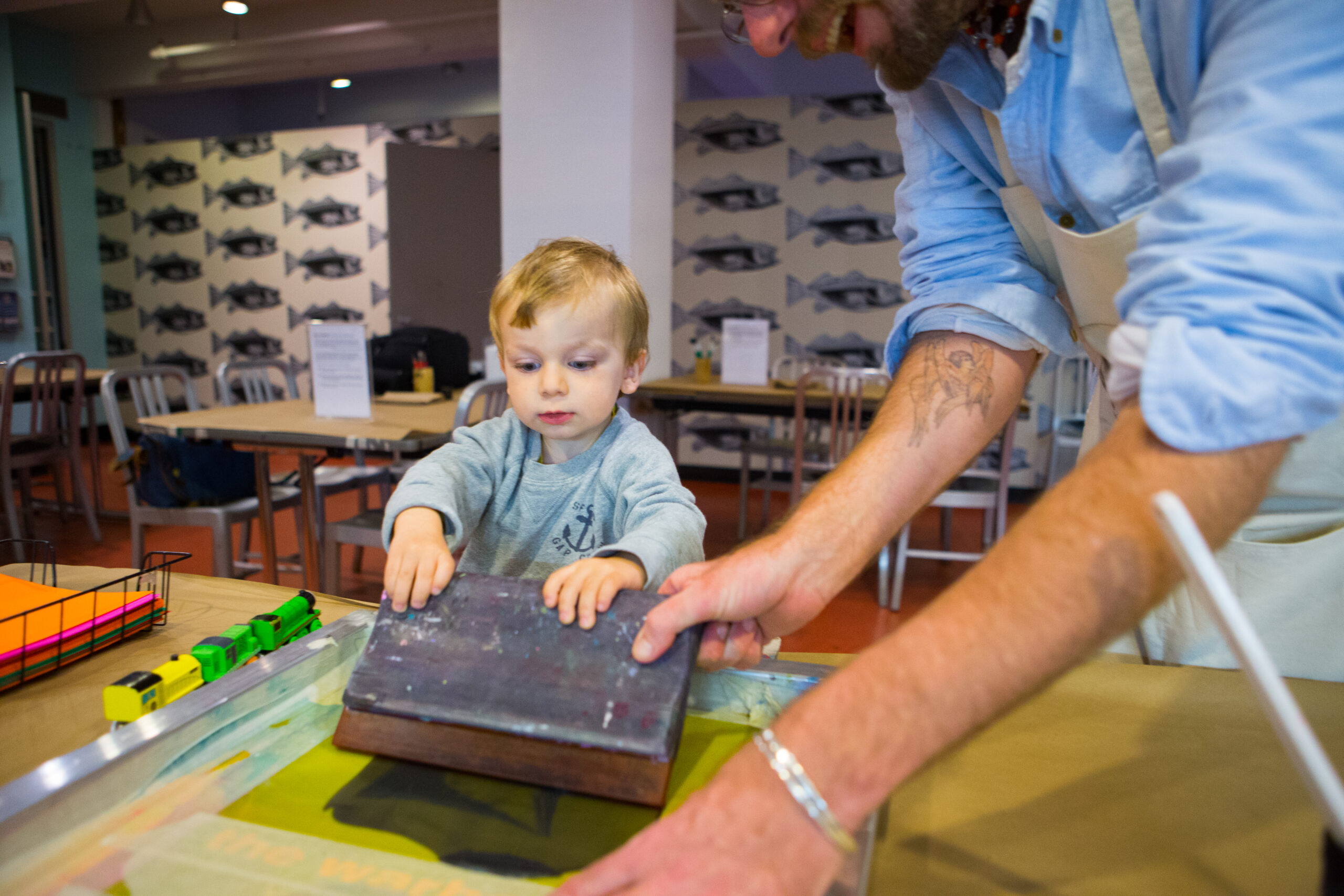 An artist educator wearing a blue button-down shirt with his sleeves rolled to his elbows and a beige apron helps a toddler with blonde hair make a screen print in the Andy Warhol Museum's Factory studio.