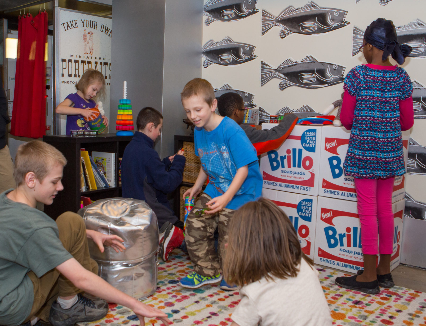 Seven children play with toys in a space with a colorful rug, Warhol Fish wallpaper, small black bookshelves, and four stacked cardboard Brillo boxes.