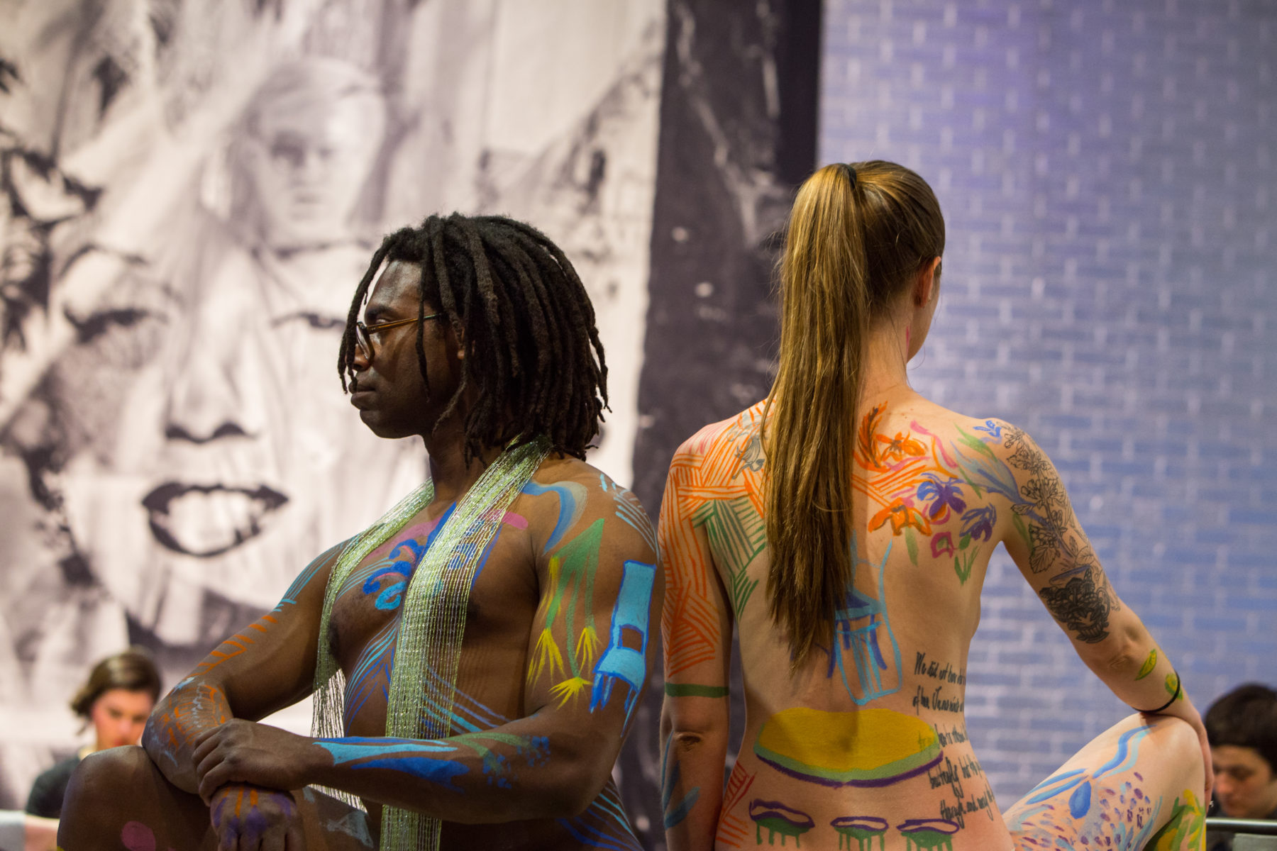 Two models posing in The Warhol entrance space. The model on the left is an African-American man, right hand resting on his propped up knee, his left hand grabbing his right wrist. He is standing back to back with a Caucasian woman, her left arm by her side and right hand grabbing her right propped up knee. Both models have a variety of small pictures painted on them with colorful body paint.