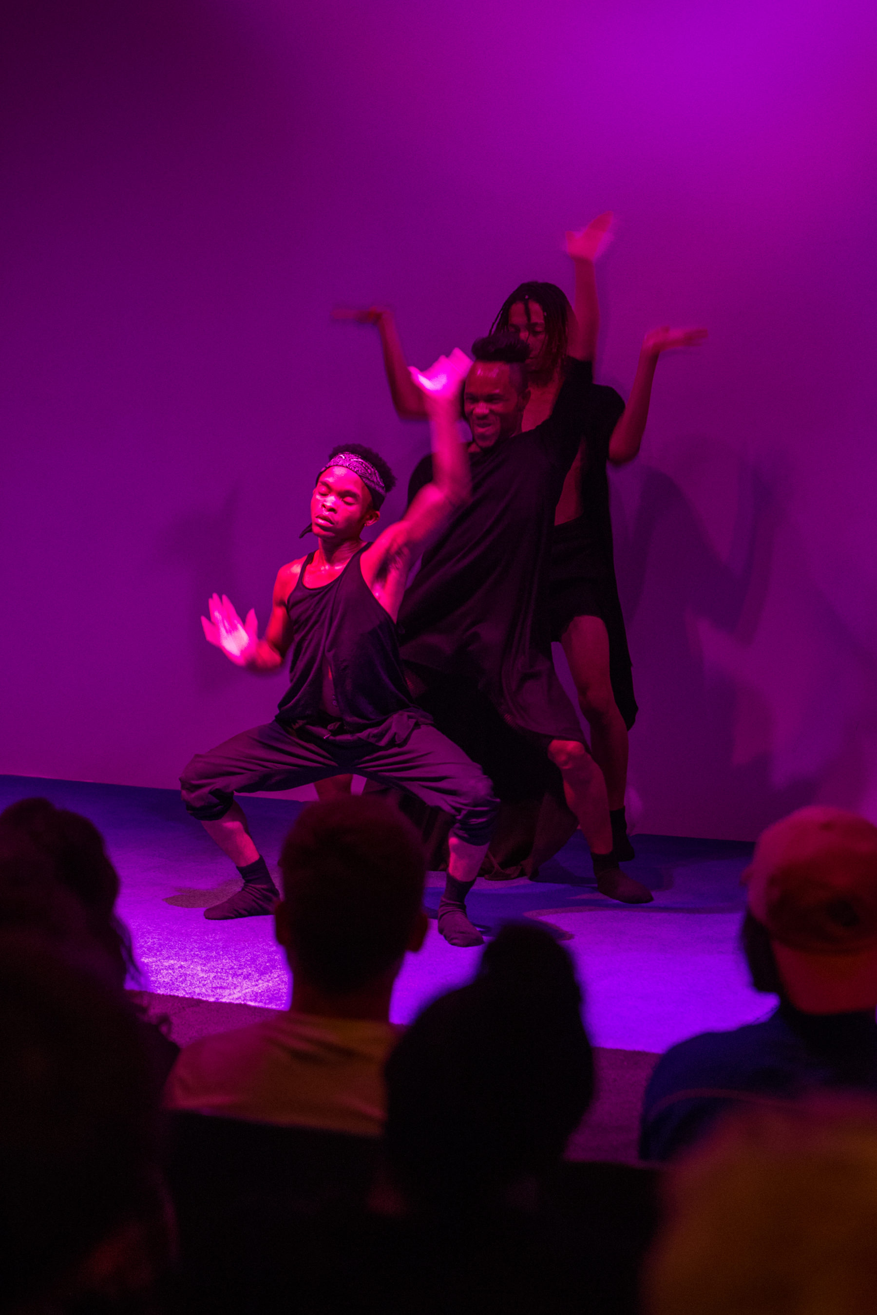 Three performers pose in a line, the performer in the front squats with his hands raised. Toward the bottom of the image we get a glimpse of audience members' heads