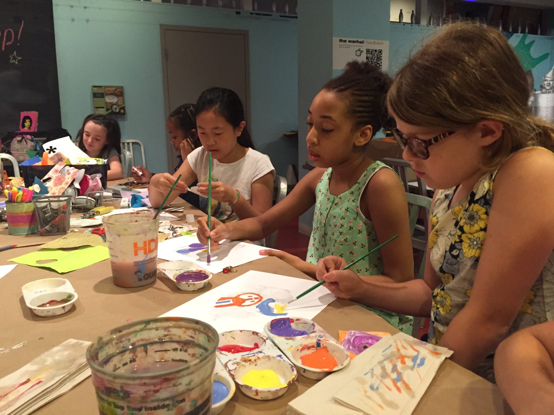 A group of girls sit at a table strewn with plastic water cups, paints, and paper as they paint.