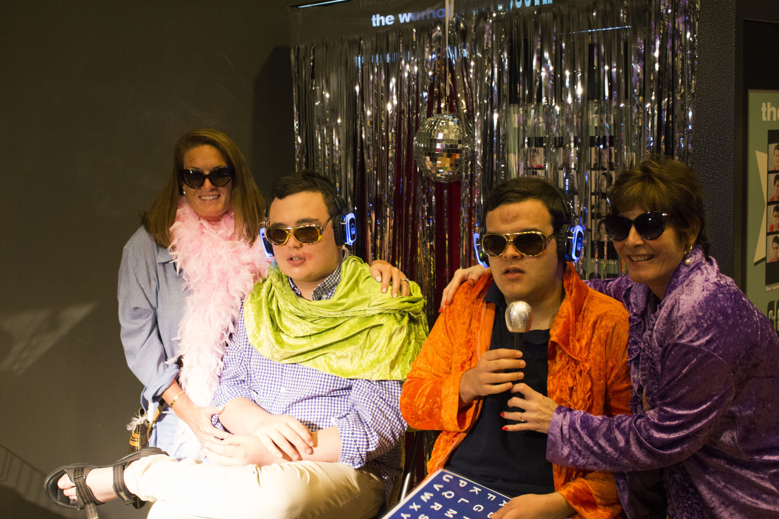 Four individuals pose for a picture against a backdrop of silver streamers and a small disco ball. On the left is a woman with brown hair, a pink feather boa, and dark sunglasses who has her arm around a man with short brown hair, sunglasses, and a green scarf. Next to them is a Man wearing sunglasses and an orange jacket. The woman on the far right wears a purple jacket and dark sunglasses, and she holds a microphone for the man at her side.