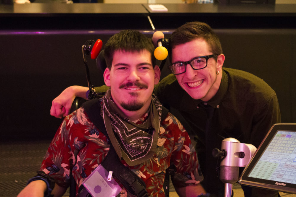 Two men pose for a picture. The man on the left has short black hair, a mustache, and a beard, and wears a bandana around his neck. He sits in a wheelchair. The man on the right has brown hair, rectangular black glasses, and has his arm around the man on the left.