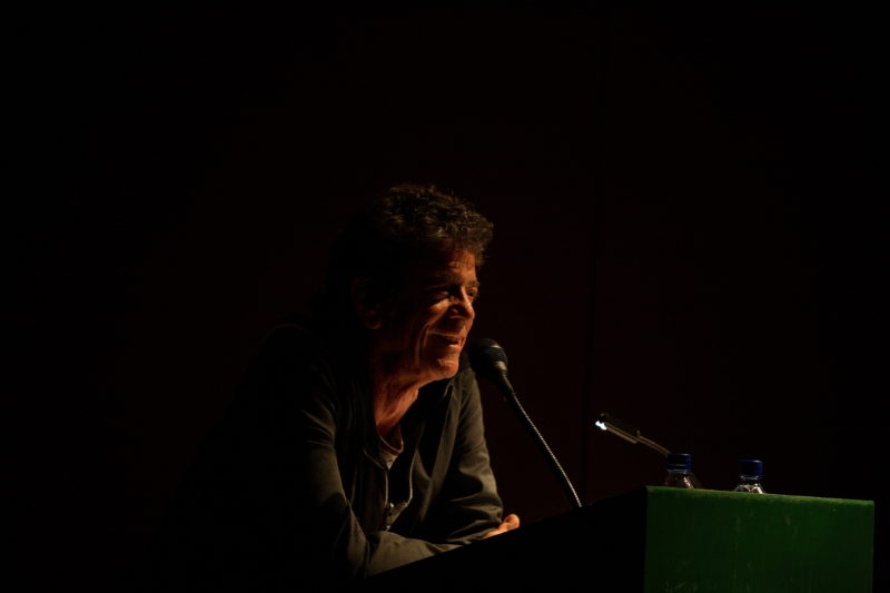 Lou Reed leans forward onto his elbows towards a microphone. His face is faintly illuminated, but most of the photograph is in shadow.