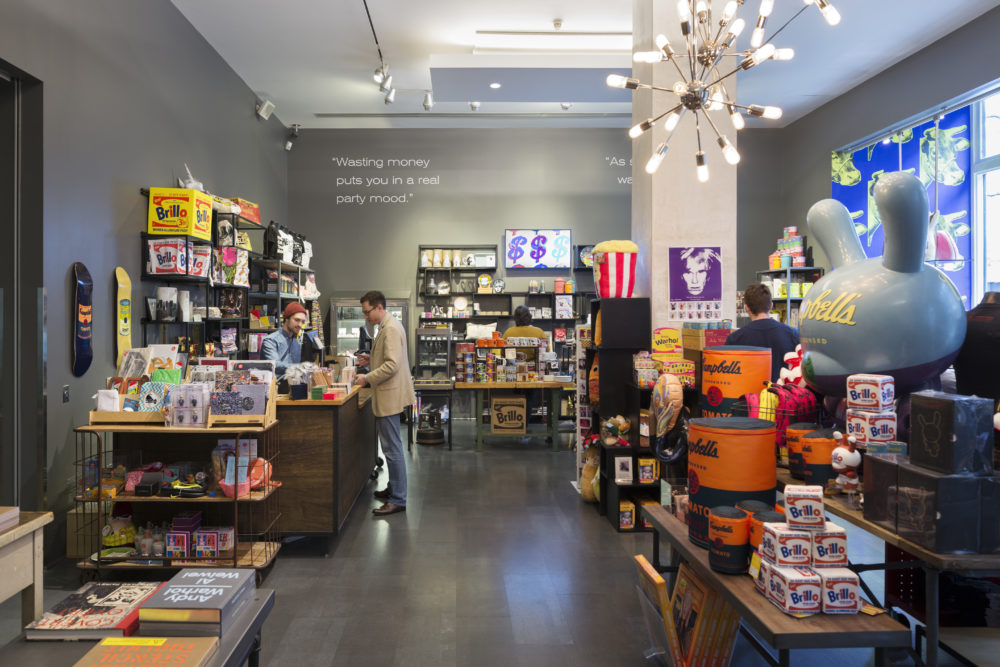 A man in a beige jacket stands at a counter in the Andy Warhol Store. The room is filled with tables and displays featuring books, soup cans, screen prints, and other warhol memorabilia. A quote painted above the shelves on the back wall reads Wasting money puts you in a real party mood.