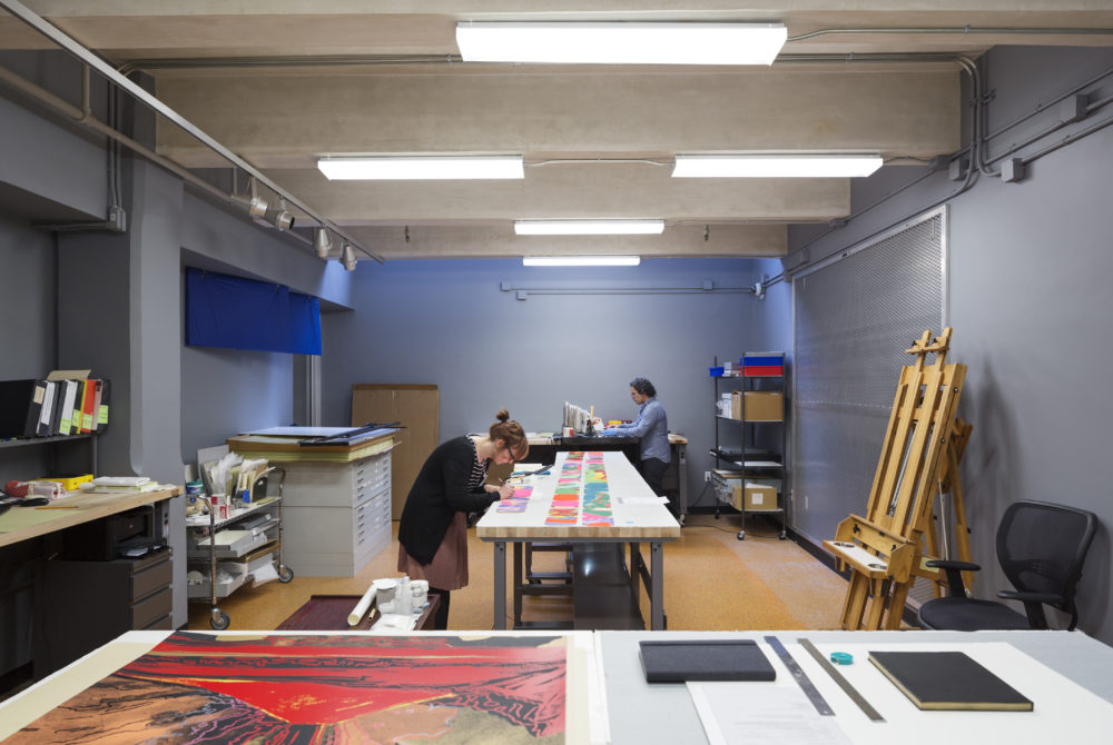 Conservationists work to preserve pieces. A woman in a beige skirt and black cardigan bends over a table in the center of the image, arranging colorful prints. In the background, a man with curly black hair and a denim shirt works at another table.