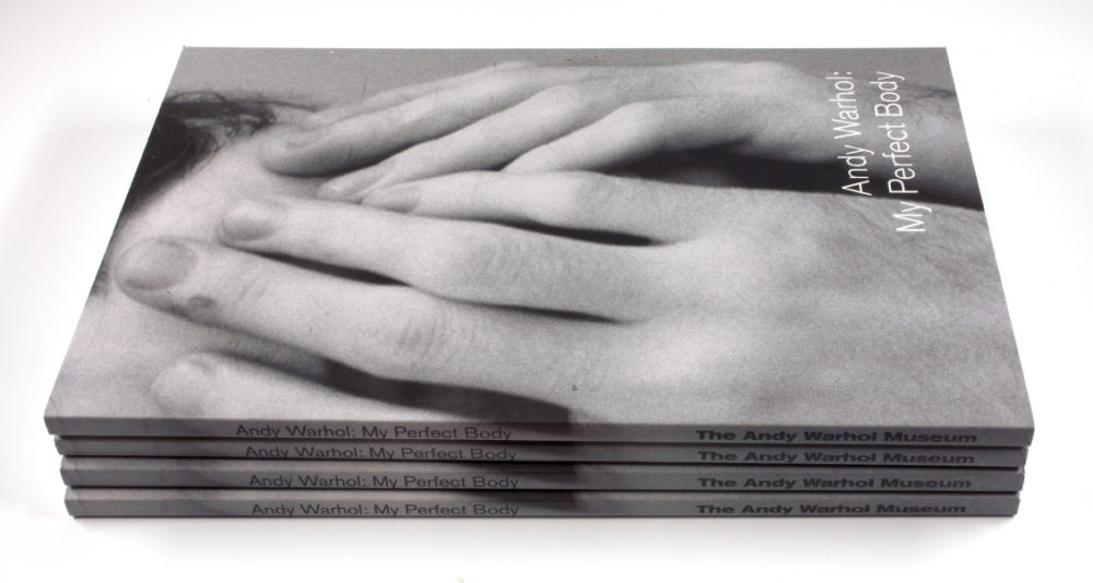 A stack of catalogues from the Andy Warhol: My Perfect Body exhibition featuring a black and white photograph of Andy Warhol with his hands covering his face.