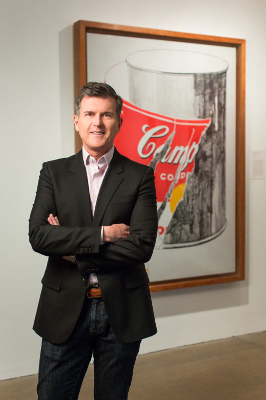 A man stands smiling with his arms folded across his chest. He is wearing dark blue jeans and a black blazer. He is standing in front of an Andy Warhol artwork showing a Campbell's soup can with a torn label.
