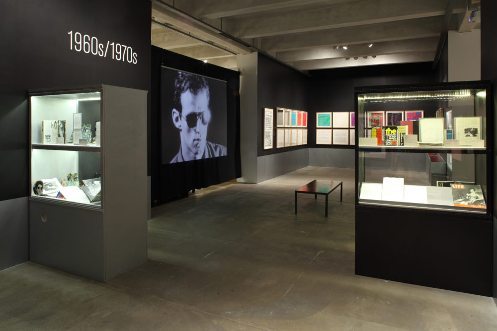 A gallery featuring a screen playing a screen test, several rows of pictures hanging on the walls, and a few glass display cases filled with archival material.