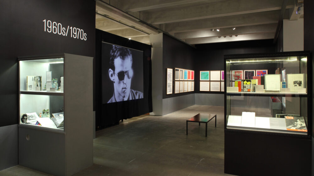 A gallery featuring a screen playing a screen test, several rows of pictures hanging on the walls, and a few glass display cases filled with archival material.