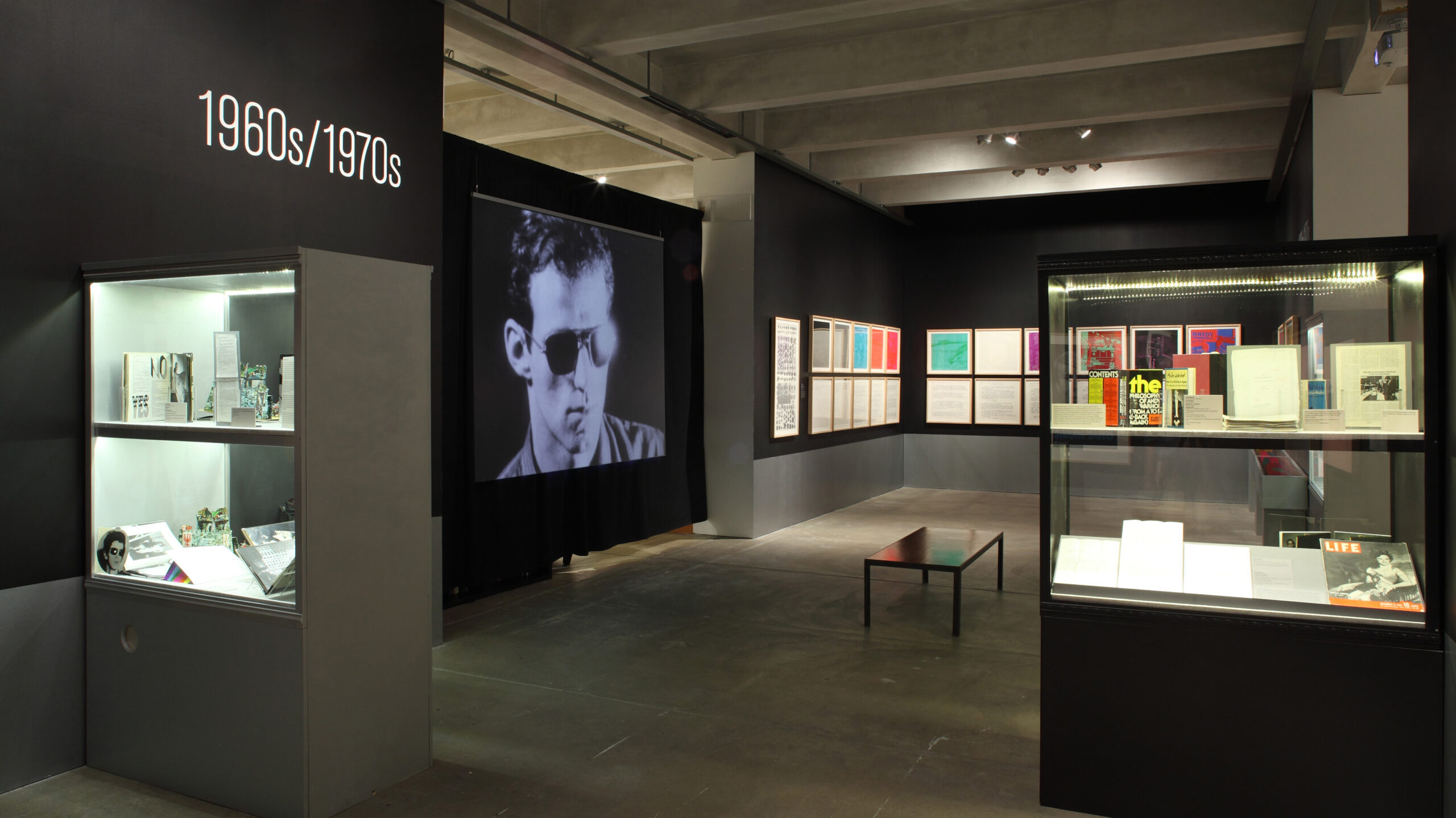A gallery featuring a screen playing a screen test, several rows of pictures hanging on the walls, and a few glass display cases filled with archival material.