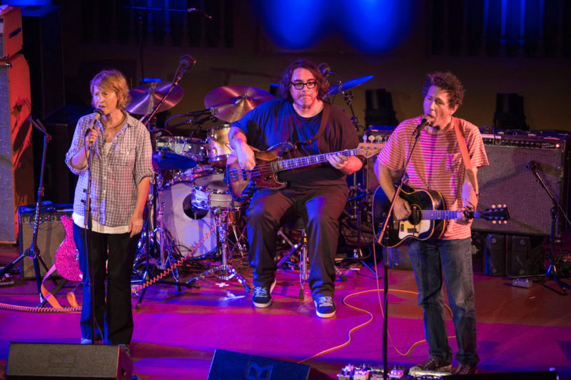 A three person musical act performs. The band member in the center sits, holding an electric guitar. To his left is an acoustic guitarist, and to his right is the vocalist. Both are standing.