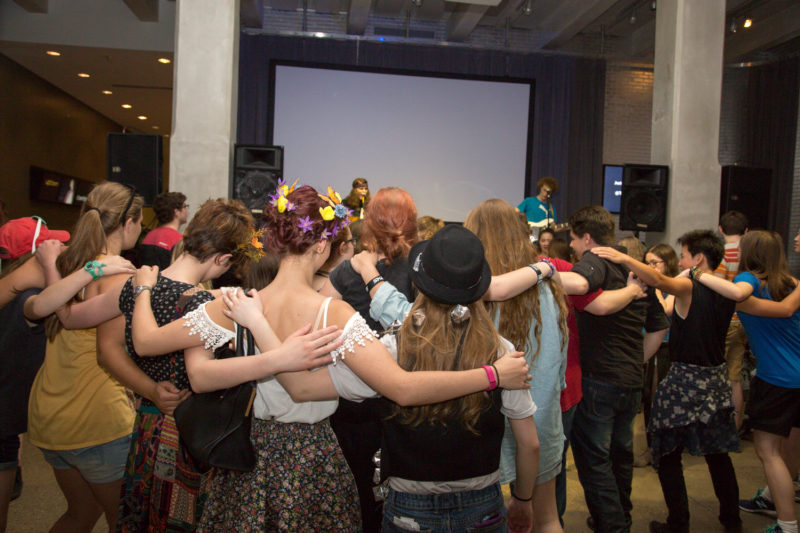 A group of teens standing side-to-side with their backs facing the viewer. They are all in an embrace, hands and arms around one another's backs.