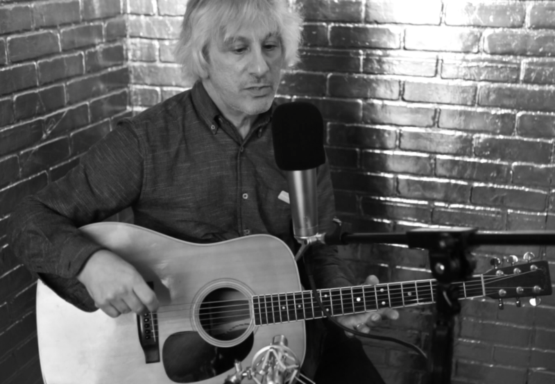 A person holding an acoustic guitar sits at a recording microphone.