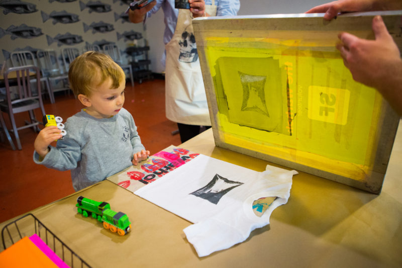 A toddler holding a yellow toy train car in his right hand looks at a white t-shirt on a table. The t-shirt has a black silkscreened image that looks like a rectangular pillow, representing one of Andy Warhol's Silver Cloud sculptures. In the upper-right-hand corner of the photograph a pain of hands holds a yellow screen with the pillow image resting on the table.