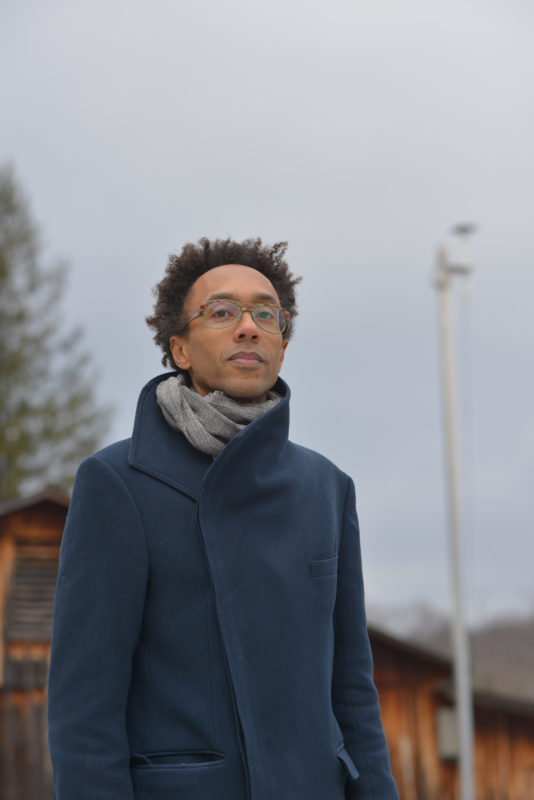 Jace Clayton wearing a blue wool coat and gray scarf standing against a gray sky.