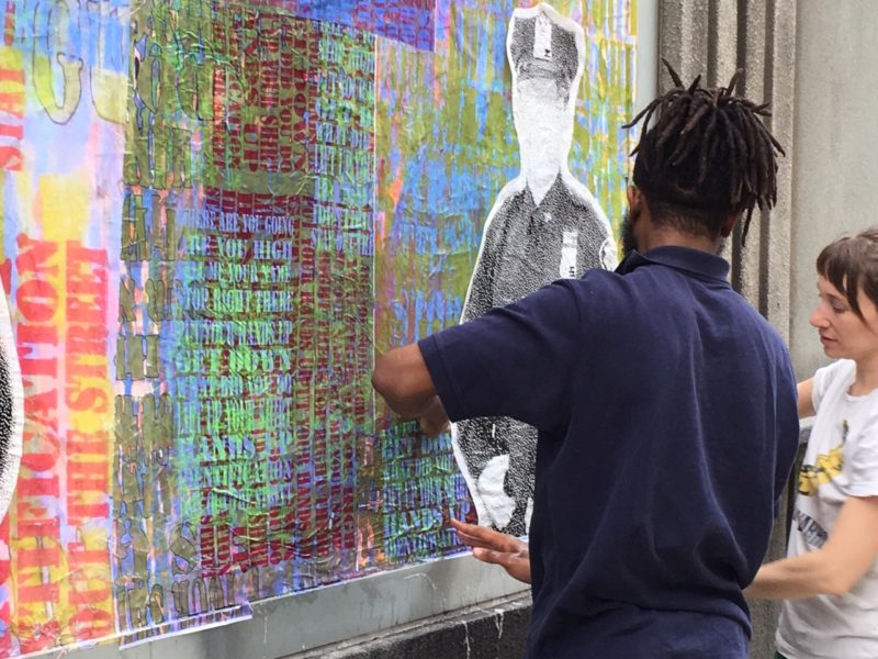 Two artists work on a large installation piece featuring overlapping colorful words and a faceless, black and white image of a police officer.