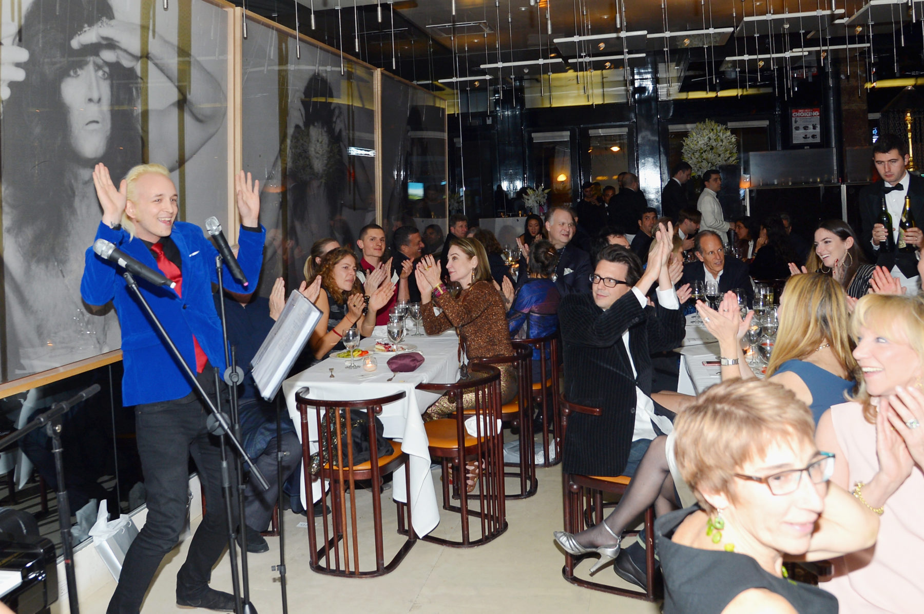 A man in a blue velvet blazer and red tie stands in front of a microphone and throws his hands in the air, smiling at a room full of applauding seated guests.