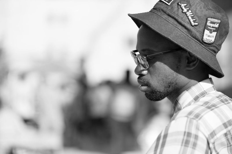 A black-and-white photo of DJ Naeem in profile, wearing a black hat adorned with patches that say Andy Warhol and Campbell's Soup.