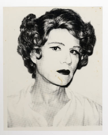A black and white photograph shows Andy Warhol from the chest up in drag, wearing a short coiffed brown wig, heavy face powder, black eyeliner, and dark red lipstick. He turns his head slightly and stares at the camera, with a loose white blouse unbuttoned at the neck.