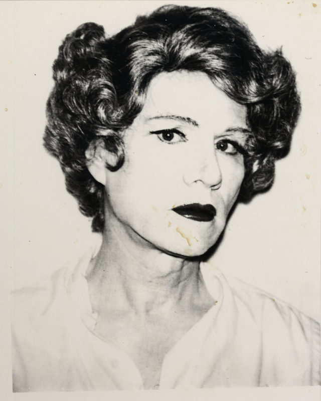 A black and white photograph shows Andy Warhol from the chest up in drag, wearing a short coiffed brown wig, heavy face powder, black eyeliner, and dark red lipstick. He turns his head slightly and stares at the camera, with a loose white blouse unbuttoned at the neck.