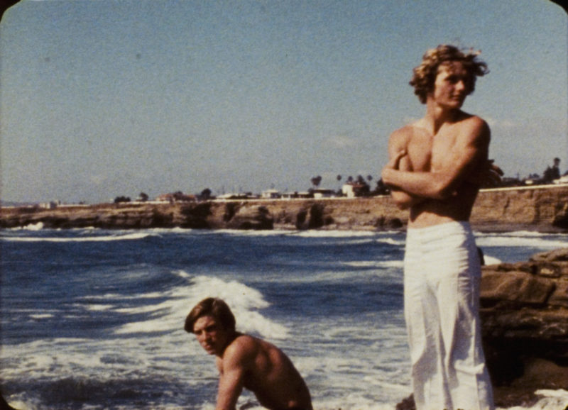 Photograph of the ocean coast with one man, shirtless and wearing white pants, standing and one man kneeling, shirtless and looking toward the viewer.