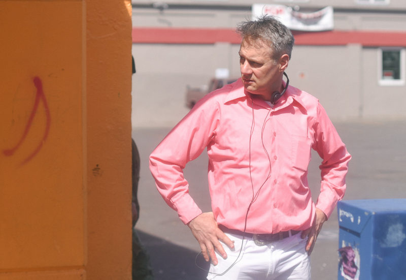 Man in a bright pink shirt and white pants stands with his hands on his hips. He is outside, standing next to a bright orange column, with black earphones around his neck.