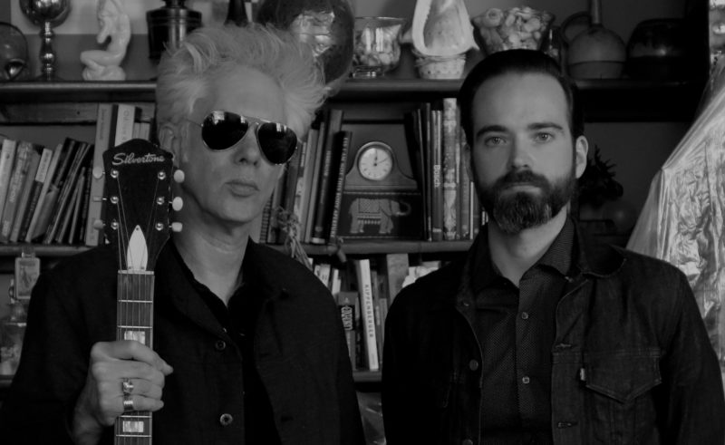 Black and white image of two men standing in front of a book shelf. The man on the left is wearing sunglasses and holding a guitar. The man on the right has a beard and is wearing a dark shirt. Both are looking directly at the viewer.