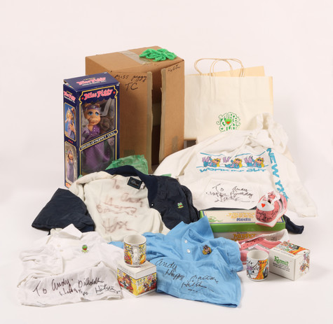 An assortment ofobjects arranged in front of a cardboard box. The objects include a Miss Piggy dress-up Muppet doll, still in its box; a shopping bag with an image of Kermit the Frog printed on it; t-shirts and polo shirts with decorated small Muppet patches and autographs addressed to “Andy”; and Miss Piggy and Kermit the Frog mugs.