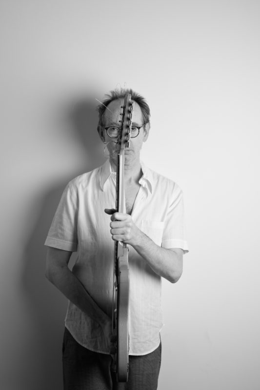 A black and white image with Arto Lindsay holding an electric guitar in front of him, the head of the guitar is in front of his face and turned sideways, the body of the guitar is in front of his torso.