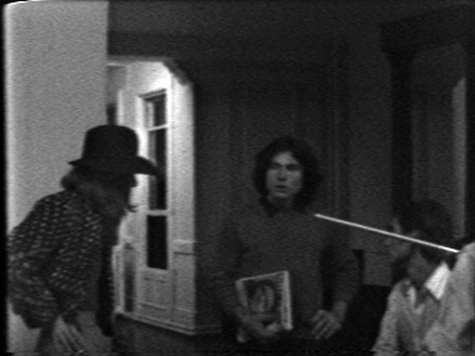 A black and white photograph of three men gathered in a room. The man on the left is in profile, a cowboy hat on his head. The man in the middle holds a portrait of a woman in his right hand and places the other on his him. The third man is seated, positioned halfway off the right hand edge of the image.