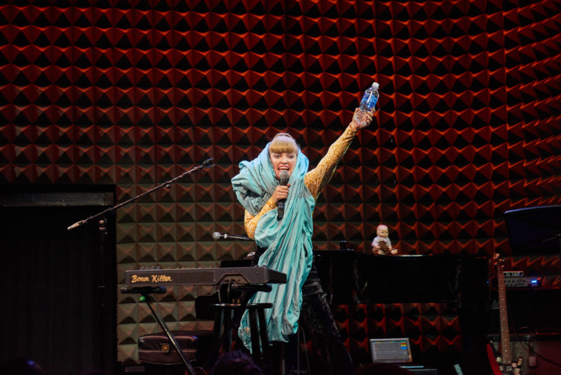 A woman with blond hair that is in a ponytail is standing on a stage with a red, textured wall in the background, talking into a microphone and holding a bottle of water in the air. She's draped in a light blue material that is wrapped around the back of her head and goes down to her knees. She's wearing a yellow shirt underneath and black pants. The stage she is standing on has various instruments on it as well as a baby doll on top of the piano.