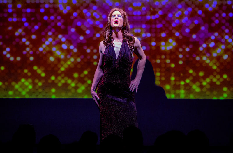 A person is on stage dressed in a red, shimmery dress. They appear to be singing. The person has long, red hair and is wearing dark, purple eye shadow and red lipstick.