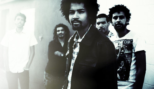 Five men standing in front of a wall. One man on the left has short, dark hair and is wearing a white shirt and dark pants. The man next to him has long dark hair and a mustache and beard. He is wearing a black shirt. The man in the middle has dark hair and a mustache and beard. He is wearing a light colored shirt with a dark colored coat over it. The next man has dark colored short hair and a mustache and beard. His body is hidden behind the others. The last man has dark colored hair and a mustache and beard. He is wearing a light colored shirt with an unrecognizable print on the front of it