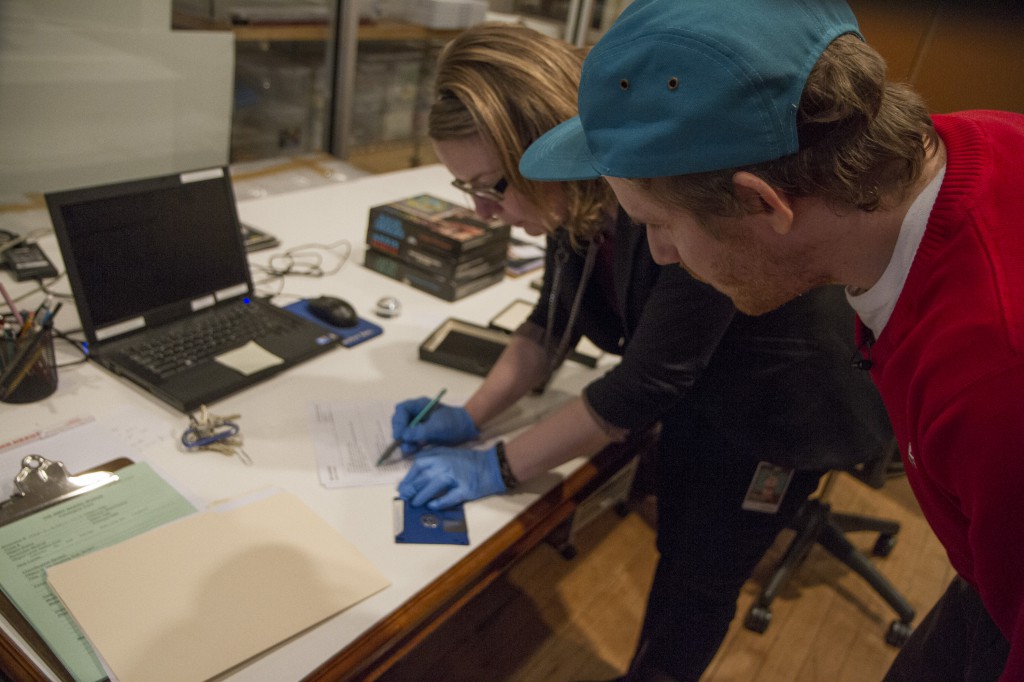 A man and a woman crouch over a desk. The woman wears blue disposable rubber gloves, holding a pencil in one hand and a floppy disk in the other. The man wears a red sweater and bright blue baseball cap and watches the woman as she takes notes.