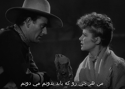 The black and white image is taken from the 1939 black and white film, Stagecoach, starring the American actor John Wayne. In the image, actors John Wayne (on the left) and Claire Trevor (on right) face one another, in conversation. Wayne’s demeanor and expression are calm as he casually leans onto the wooden fence standing in between the two of them. He loosely wears a wide-brimmed, tan cowboy hat and a dark jacket. On the right, Trevor leans in towards Wayne. Her attitude appears serious—her brows are wrinkled and her hands are held closely together resting on the fence. Though the background is out of focus, the dim outline of tree branches suggests that the two are outside. At the bottom of the image there are subtitles in Farsi translating the dialogue. Translation is not available.